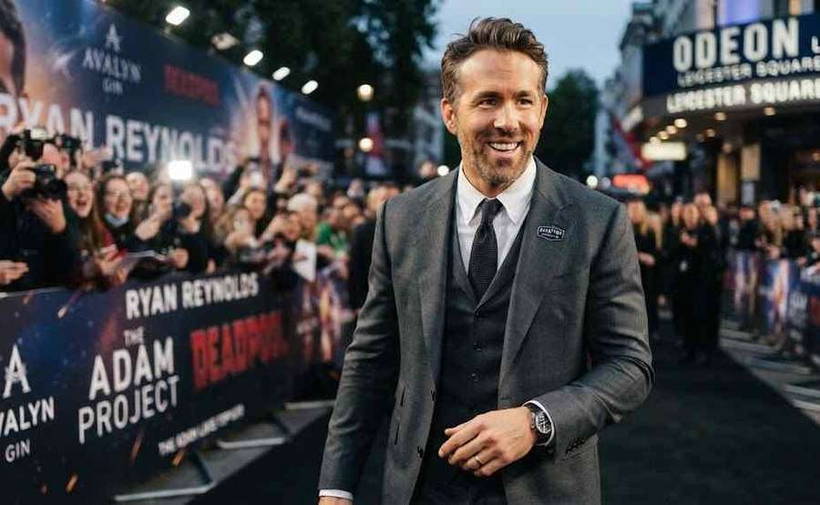 Ryan Reynolds smiling on a crowded red carpet event in a stylish grey suit.